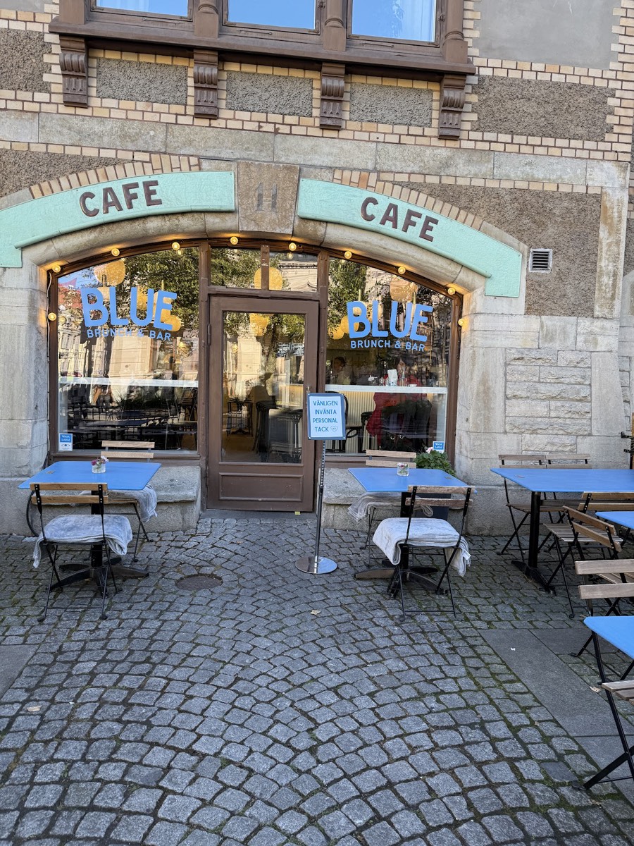 Blues fasad på Vasagatan 11: turkos Café-skylt i stenbågen, BLUE i fönstren och blå bord på trottoaren